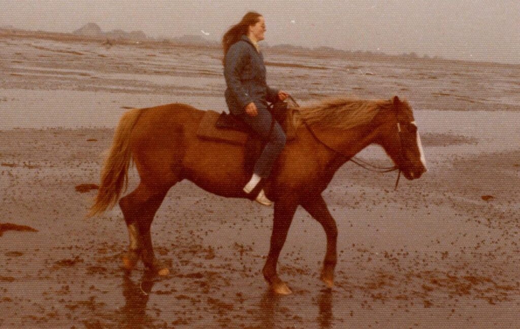 A young woman in a blue jacket is riding a horse in the wind on a beach with large ocean waves crashing in the background.