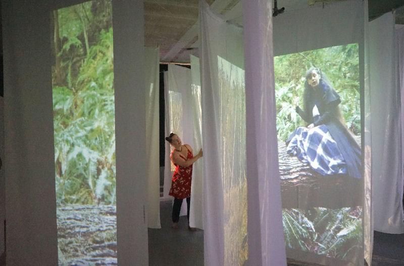 A still photo of artist Emily van Lidth de Jeude's first what dot home installation in Amsterdam. A room is filled with hanging white fabric panels, on which is projected a fern-covered hillside and a big log, ontop of which a woman is sitting expressively speaking. Emily is walking between the panels of fabric, reaching around and pushing one panel to the side as she passes it.