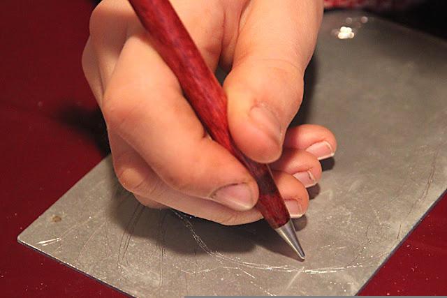 A hand holding a metal scribe, scratching a design into a metal plate.