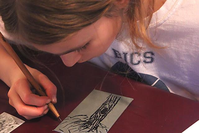 A child leaning over a metal plate with a tree drawn onto it, and scratching the design into the plate with a metal scribe.