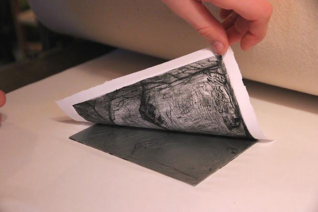 A hand is pulling back a piece of paper from the pressed etching plate, and the printed image is clearly visible on the paper. The image appears to be a simple drawing of trees and mountains.