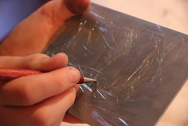 An image of a hand holding a metal scribe and scratching a drawing into a metal plate.