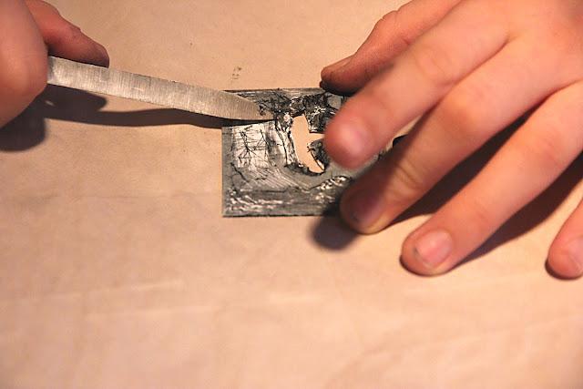 And image of a hand holding a metal plate with a drawing and a hole in the middle, and a hand scraping the plate with a sharp-edged tool.