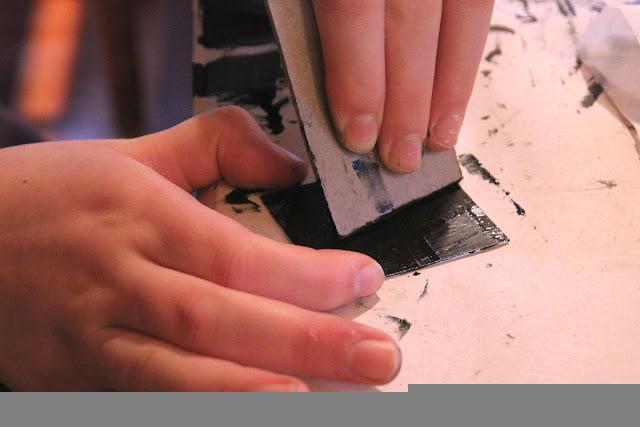 One hand holds a metal plate stable on a table while another hand smears wet black ink into the plate using a square of cardboard.