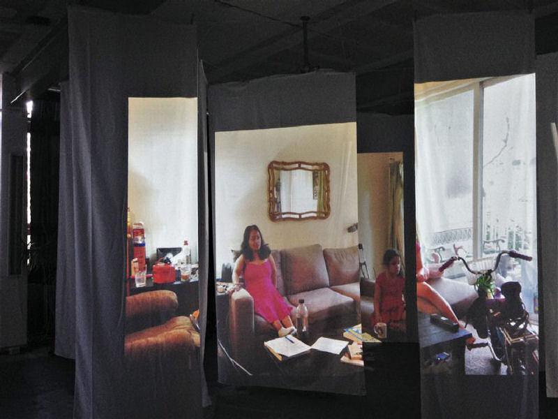 A photo of Emily van Lidth de Jeude's video installation, what dot home. A series of fabric panels hang loosely in the dark, and an image is projected onto them of a very short Mexian woman, sitting on a couch with a mirror above her. Around her can be seen an easy chair, medications, small crutches, a coffee table with papers strewn over it, a child's bicycle, and a young black/indigenous child. Both are wearing pink dresses.