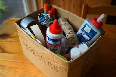 A cardboard box labelled "adhesives", filled with bottles of glue, glue-sticks, and rolls of tape.