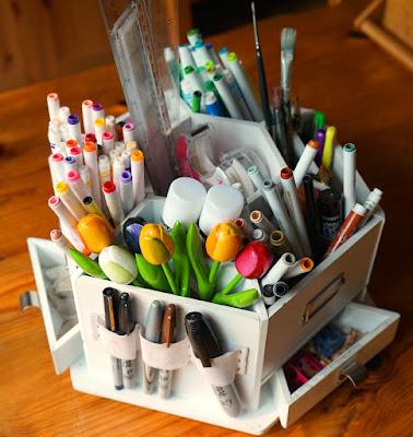 A white hexagonal art caddy filled with coloured felt pens, glue sticks, rulers, tape, paint brushes, pencils, permanent pens, erasers, and a drawer with pencil sharpeners and shavings.