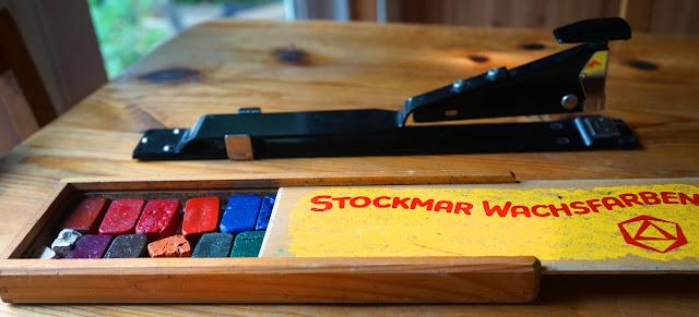 A box of Stockmar-brand brick-shaped beeswax crayons, and a long-reach booklet stapler behind it.