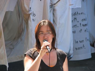 A woman with orange-brown straight long hair stands in front of painted sheets, singing into a corded microphone that she's holding up with her right hand. She is wearing a black t-shirt, a black choker-necklace with an orange stone on it, and a pair of glasses.