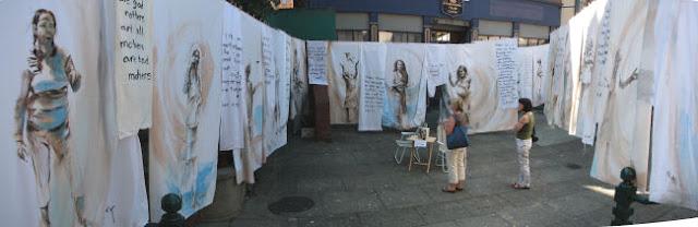 A concrete-tiled city street is filled with clotheslines, from which hang numerous tattered and stained white bed sheets. Each sheet is painted with a portrait of a mother, speaking or emoting. Between the portraits are hung smaller pieces of fabric, covered in hand-painted text. In the middle of the image, two women are standing, arms folded, looking at the sheets. A small table is beside them, with a book on it, and two lawn chairs.