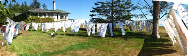 In a large open grassy yard with two large trees and a large old character home, multiple clotheslines support dozens of white tattered sheets, blowing in the wind. The sky is clear and it's a sunny day. It's difficult to see because the image is small, and taken from a distance, but the sheets are painted with portraits of mothers, and words.