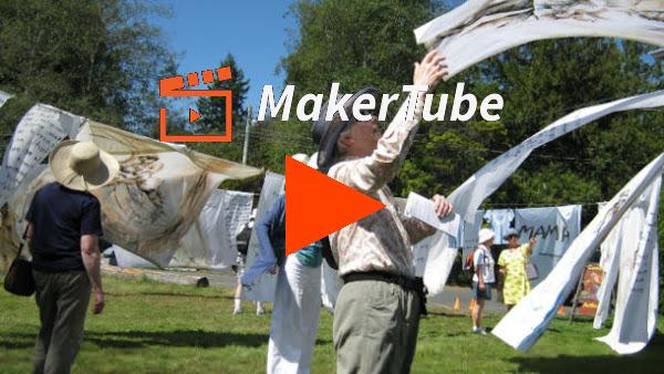 A video preview image (clickable link). An image of multiple people standing around between dozens of hanging sheets, all painted with portraits of mothers, and blowing in the wind. The woman at the forefront of the image is reaching up to catch a sheet. It's a sunny day on a large lawn, and all of the people are wearing sun hats. The image also has the MakerTube logo and a big orange PLAY button on it.