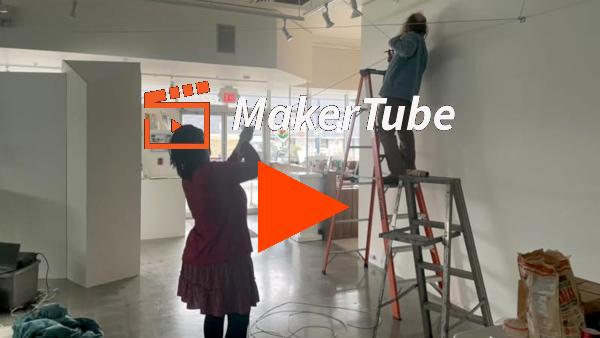 A video play link. An image of two people in a gallery, hanging up cables. There is an orange and white Maker Tube logo and a big orange play button superimposed on the image.