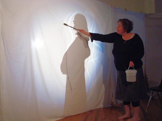 A white sheet is hung across the room with wooden clothespins, and it's lit from behind, where the shadow of a singing pregnant woman is cast against it. In front of the white sheet, a woman in a black dress stands painting with water around the shadow of the pregnant woman. The painted lines make the sheet more transparent, accentuating and ornamenting the view of the pregnant woman singing.