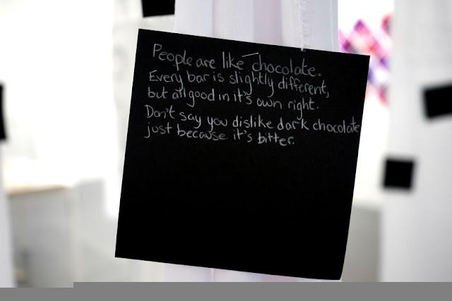 Text written by a child in white pencil on a black square card, hanging from a thread in a gallery installation. The text says: "People are like chocolate. Every bar is slightly different, but all good in it's own right. Don't say you dislike dark chocolate just because it's bitter."