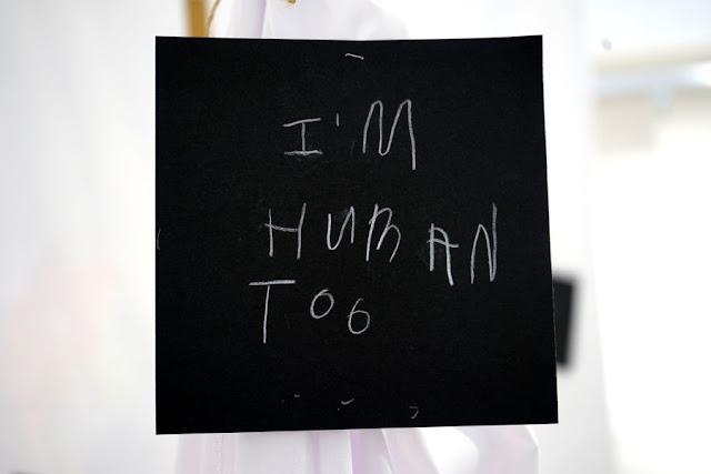 Text written by a child in white pencil on a black square card, hanging from a thread in a gallery installation. The text says: "I'm human too."
