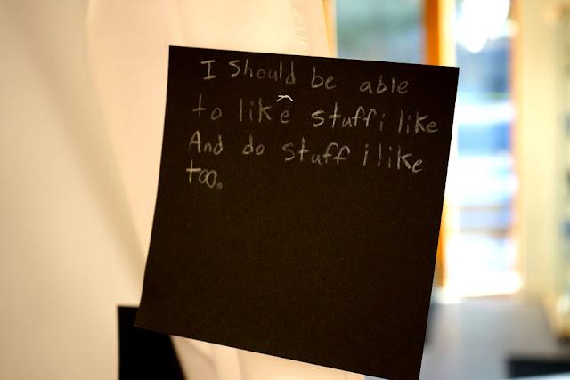 Text written by a child in white pencil on a black square card, hanging from a thread in a gallery installation. The text says: "I should be able to like stuff i like and do stuff i like too."
