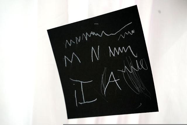 Text written by a child in white pencil on a black square card, hanging from a thread in a gallery installation. The text is not legible, but might say, "I am..."