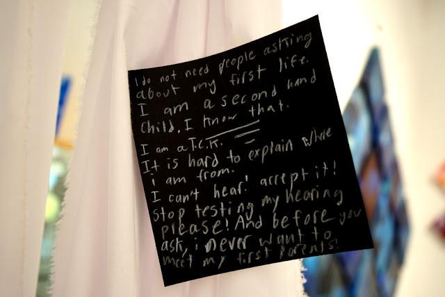Text written by a child in white pencil on a black square card, hanging from a thread in a gallery installation. The text says: "I do not need people asking about my first life. I am a second hand child. I KNOW THAT. I am a T.C.K. It is hard to explain where I am from. I can't hear! Accept it! Stop testing my hearing please! And before you ask, I never want to meet my first parents!"