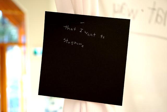 Text written by a child in white pencil on a black square card, hanging from a thread in a gallery installation. The text says: "That I want to stay home."
