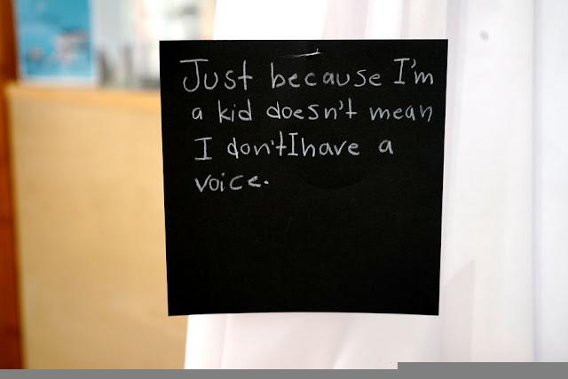 Text written by a child in white pencil on a black square card, hanging from a thread in a gallery installation. The text says: "Just because I'm a kid doesn't mean I don't have a voice."