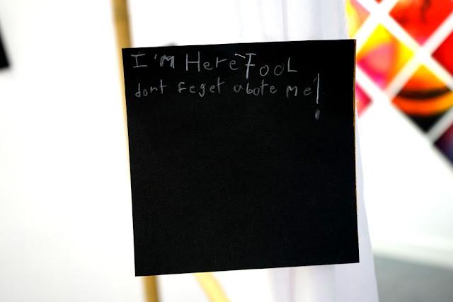 Text written by a child in white pencil on a black square card, hanging from a thread in a gallery installation. The text says: "I'm here, fool! Don't forget about me!"