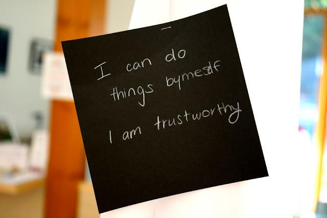 Text written by a child in white pencil on a black square card, hanging from a thread in a gallery installation. The text says: "I can do things by myself. I am trustworthy."