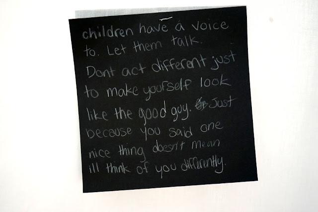 Text written by a child in white pencil on a black square card, hanging from a thread in a gallery installation. The text says: "Children have a voice. Let them talk. Don't act different just to make yourself look like the good guy. Just because you said one thing doesn't mean I'll think of you differently."