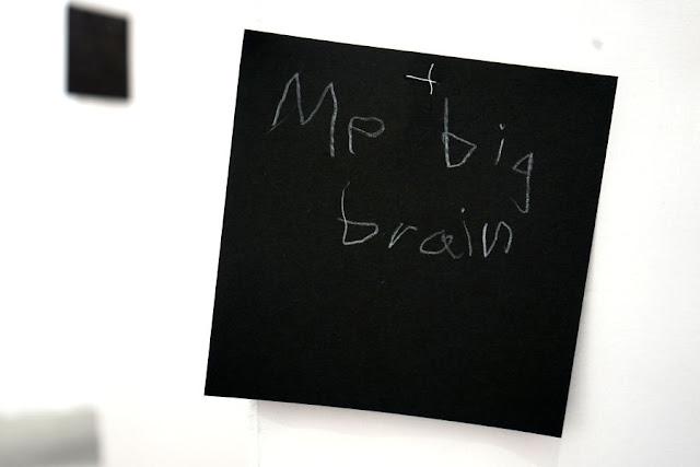Text written by a very young child in white pencil on a black square card, hanging from a thread in a gallery installation. The text says: "Me big brain."