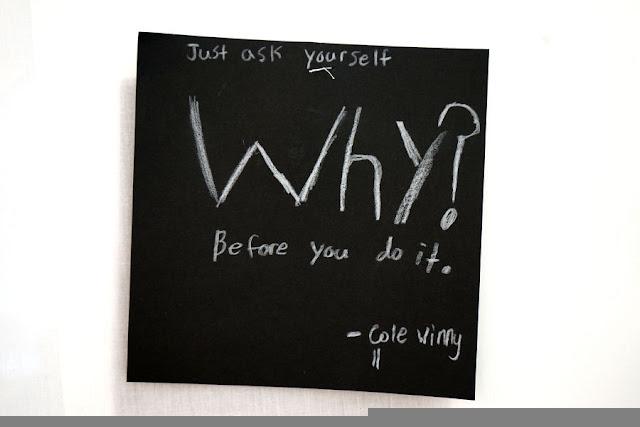 Text written by a child in white pencil on a black square card, hanging from a thread in a gallery installation. The text says: "Just ask yourself WHY before you do it."