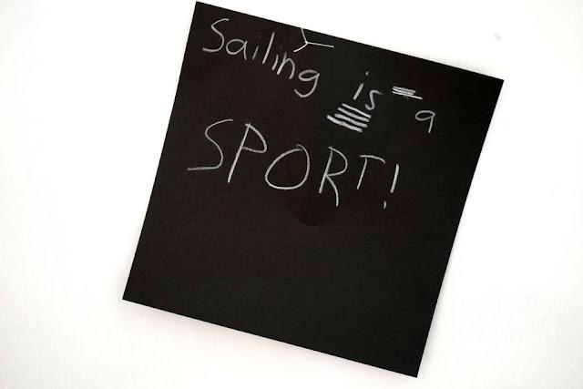 Text written by a child in white pencil on a black square card, hanging from a thread in a gallery installation. The text says: "Sailing is a sport!"
