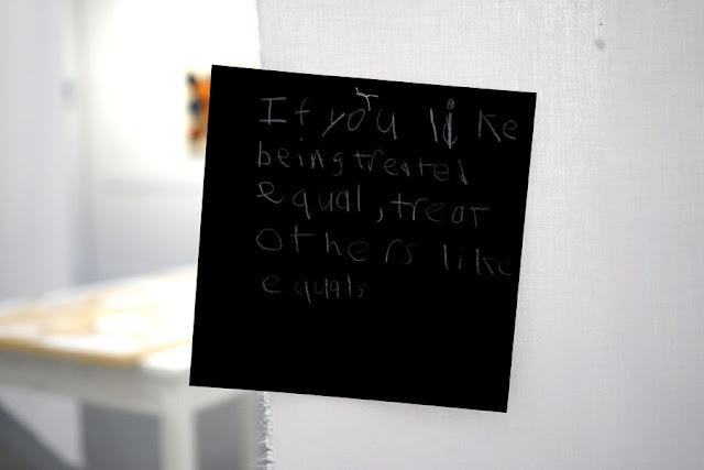 Text written by a child in white pencil on a black square card, hanging from a thread in a gallery installation. The text says: "If you like being treated equal, treat others like equals."