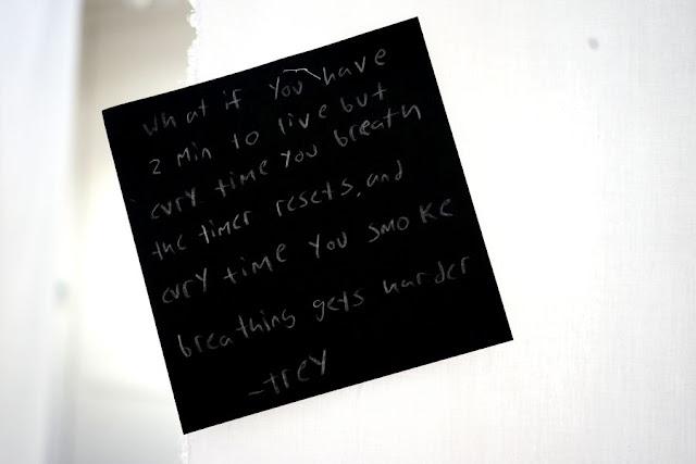 Text written by a child in white pencil on a black square card, hanging from a thread in a gallery installation. The text says: "What if you have two minutes to live but every time you breathe the timer resets and every time you smoke breathing gets harder?"