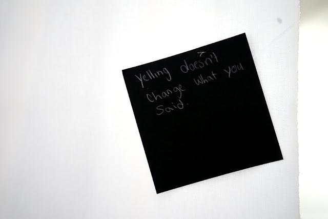 Text written by a child in white pencil on a black square card, hanging from a thread in a gallery installation. The text says: "Yelling doesn't change what you said."