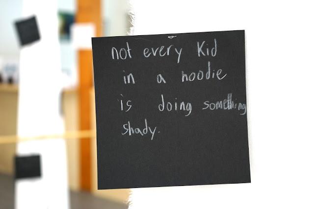 Text written by a child in white pencil on a black square card, hanging from a thread in a gallery installation. The text says: "Not every kid in a hoodie is doing something shady."