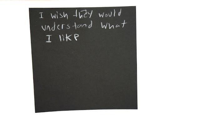 Text written by a child in white pencil on a black square card, hanging from a thread in a gallery installation. The text says: "I wish they would understand what I like."
