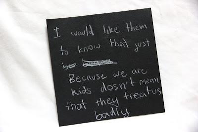 Text written by a teen in white pencil on a black square card, hanging from a thread in a gallery installation. The text says: "I would like them to know that just because we are kids doesn't mean that they treat us badly."