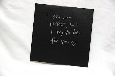 Text written by a teen in white pencil on a black square card, hanging from a thread in a gallery installation. The text says: "I'm not perfect, but I try to be, for you." There is a heart after the text.