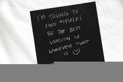Text written by a teen in white pencil on a black square card, hanging from a thread in a gallery installation. The text says: "I'm trying to find myself and be the best version of whoever that is."