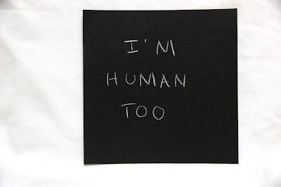 Text written by a teen in white pencil on a black square card, hanging from a thread in a gallery installation. The text says: "I'm human too."