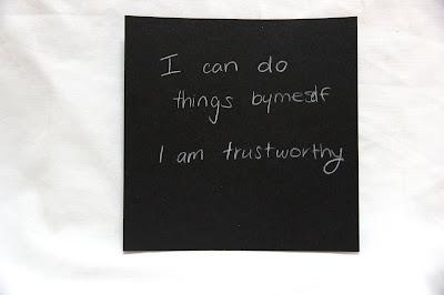 Text written by a teen in white pencil on a black square card, hanging from a thread in a gallery installation. The text says: "I can do things by myself. I am trustworthy."