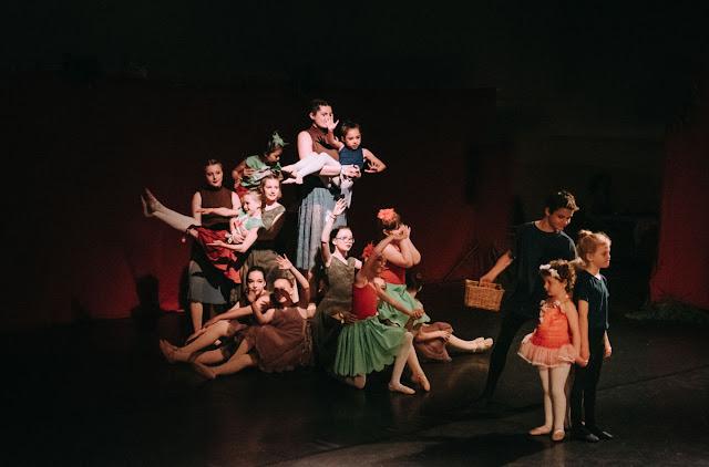 A collection of dancers in brown and green leotards and flowy skirts stands smiling and gesturing. They represent an apple tree. An older boy dressed in a black t-shirt and leggings holds a basket in one hand, and looks towards three younger dancers on the right side of the stage: A young oy also dressed in black, and two very young ballerinas, in pinkish-orange tutus.