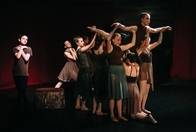 A group of dancers in brown and green leotards and flowy skirts lift the oldest dancer along, where she has fallen from the stump she initially stood on. They represent an apple tree, falling.
An older boy dancer in a brown shirt and black leggings stands off to the left, his hands crossed over his chest, looking solemnly in the direction of the tree.