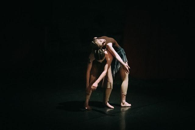 Two contemporary dancers in brown leotards and flowy green skirts stand centre-stage. One leans forward, while the other one leans over her, back-to-back, in an expression of sorrow and exhaustion.