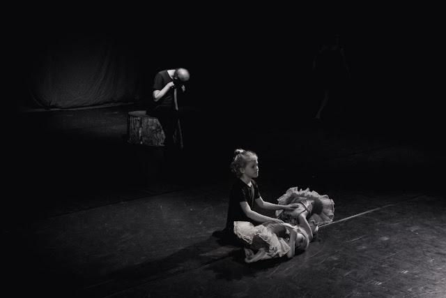 A black and white image of an old man sitting on a stump, leaning on a crutch. In front of him, a young dancer dressed in black kneels on the ground, his hands resting on the backs of two very young ballerinas in tutus, curled up on the floor, in front of him.