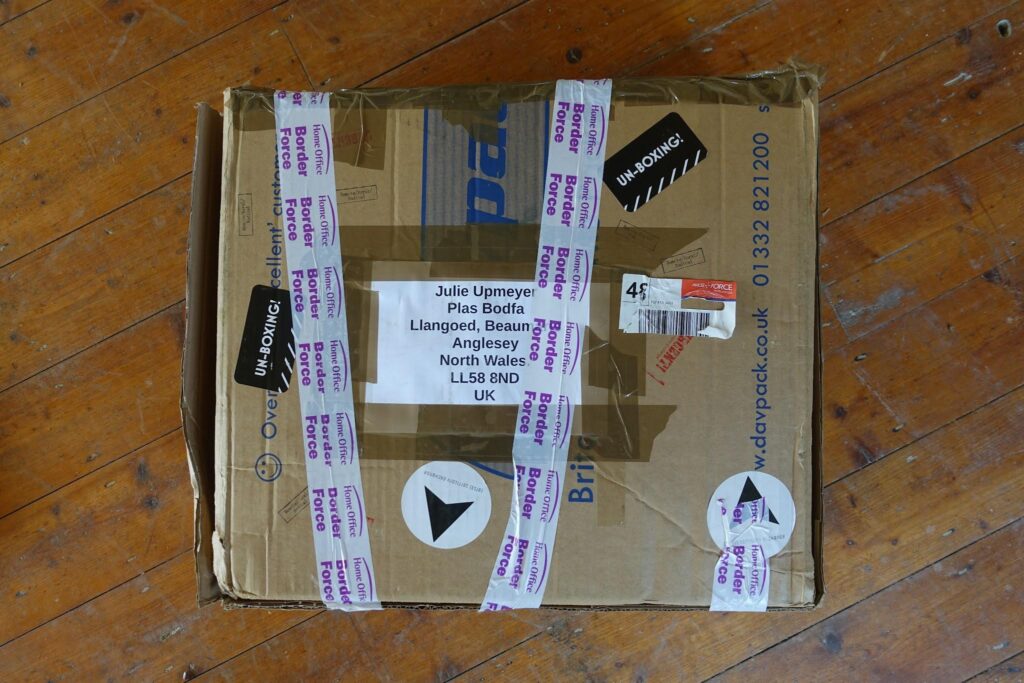A tape- and sticker-covered brown cardboard box sits on a wooden floor. Some of the stickers say Un-Boxing! and the tape says Border Force. The box is addressed to Julie Upmeyer, Plas Bodfa, Anglesey, North Wales, UK.