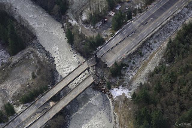 A bridge on the Coquihalla highway is shown from above, in an aerial photograph by Douglas Noblet. The two lanes of the highway break away and are seen crumbled in the river, below. A small road running alongside the highway has also been washed out by a flood coming down the mountain, into the river.