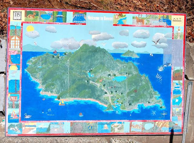 A large plywood mural is almost square, with a large painting of a green, tree-covered island, with beaches, houses, lakes, and a few sailboats, sea mammals, and birds in the blue water around it. The frame of the painting is covered with a series of small paintings of landscapes, seascapes, and other wilderness.