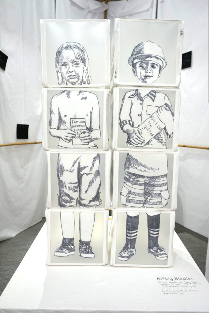 Two stacks of 4 translucent storage cubes with white fabric edges, standing side-by-side. Each stack forms a black-and-white hand-drawn portrait of a child (feet on the bottom cube, upper legs above, torso above that, and heads at the top). The children are holding papers that say "I am more sensitive than I look", and "don't call me gifted".