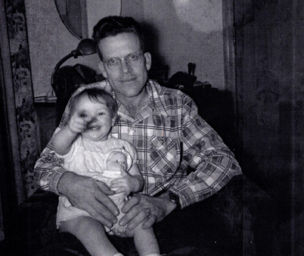 A man with short stand-up brown hair in a plaid shirt sits holding a baby on his lap, and she's holding a dolly on her own lap. He is smiling at the camera; she is laughing and pointing at the camera. Wearing a little white baby dress.